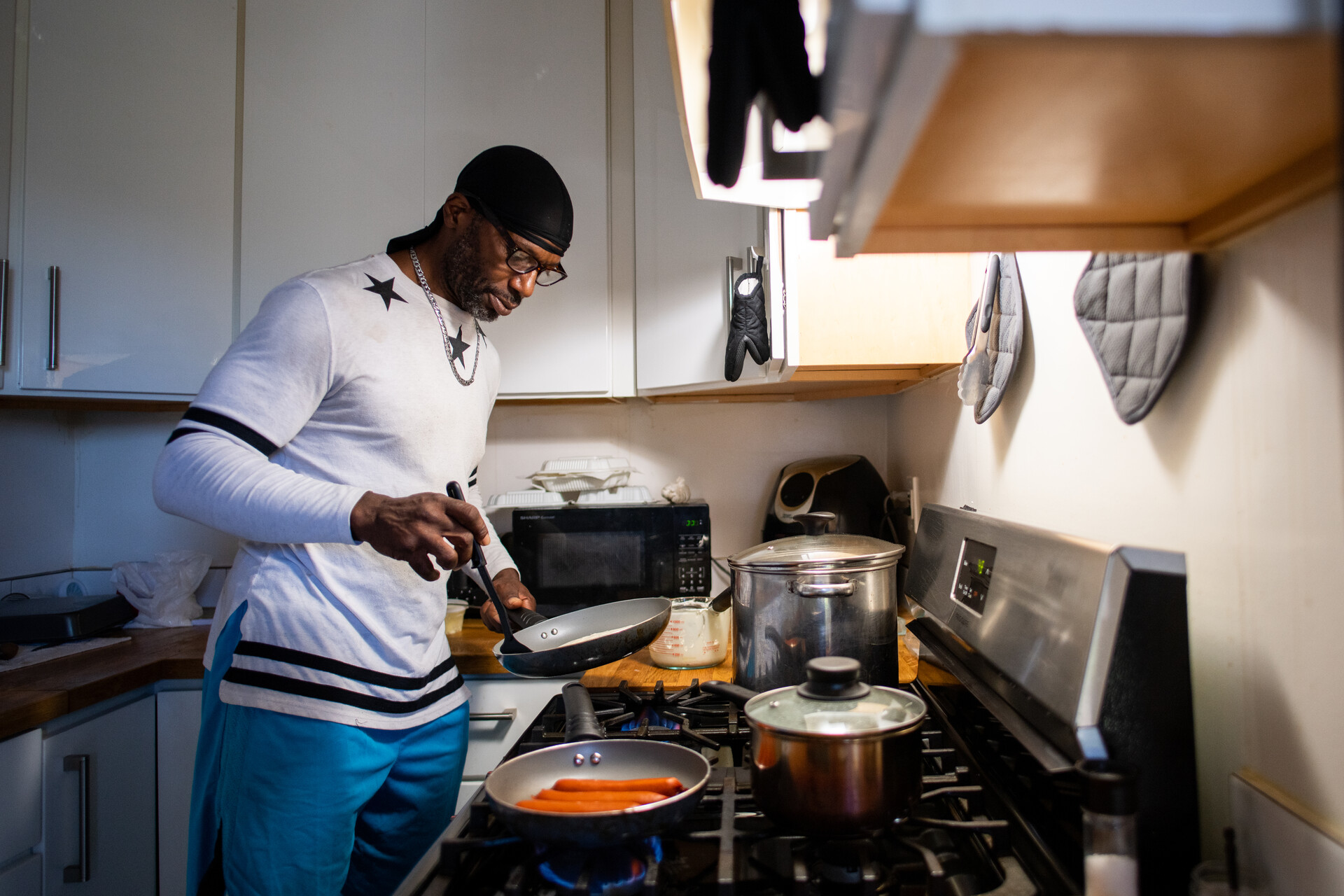 a Black man in a white shirt makes pancakes, standing at the stove in his apartment