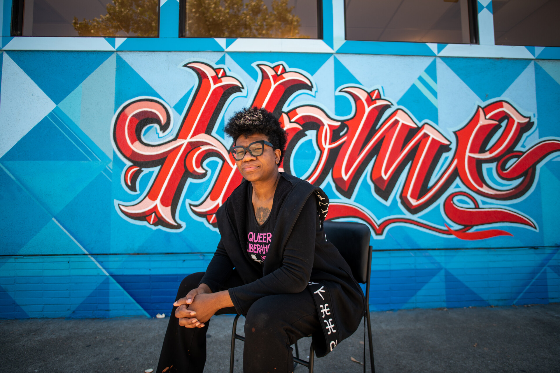 A black woman wearing a black suit and glasses sits in front of a blue and red mural that reads 