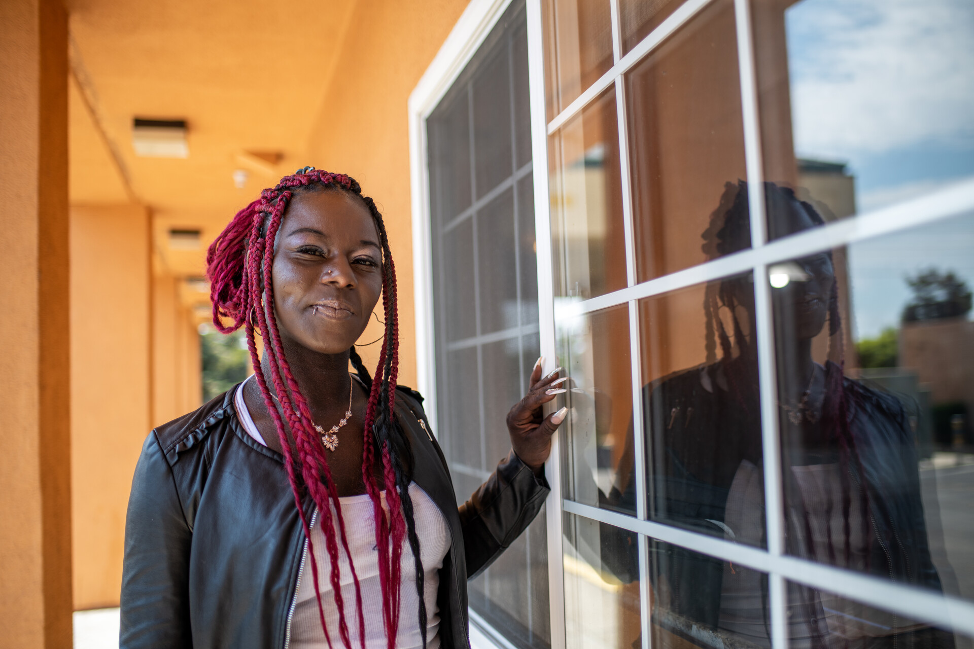 A smiling Black woman with bright red braids stands against a window and the yellow wall of a hotel.
