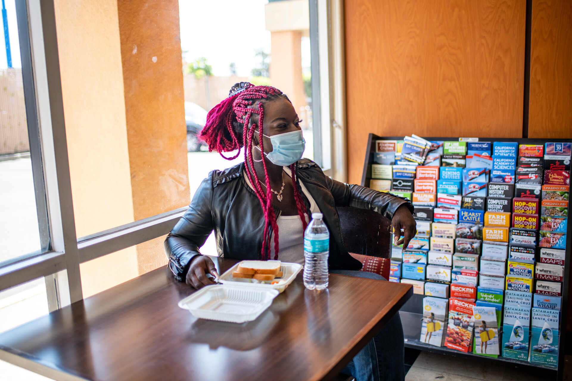 a Black woman with bright red braids eats takeout lunch at a table in a motel lobby in front of an orange wall and a display case full of pamphlets