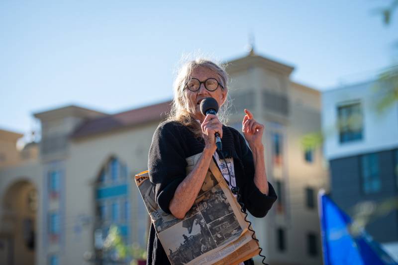 An older woman with glasses stands with a microphone with the backdrop of a building and blue sky