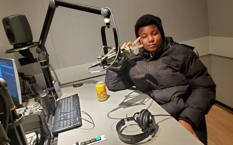 A young black woman wearing a black parka sits in an audio studio, a microphone pointed toward her mouth and headphones on the table in front of her. She holds two fingers up, pointing them toward her face. A tangerine-flavored La Croix fizzy water is on the desk in front of her.