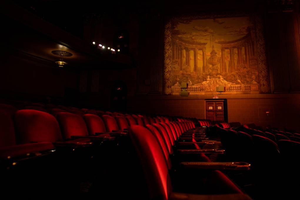 A dimly lit row of red velvet seats stretch away from the camera, leading towards an Art Deco painting or mural on the wall.