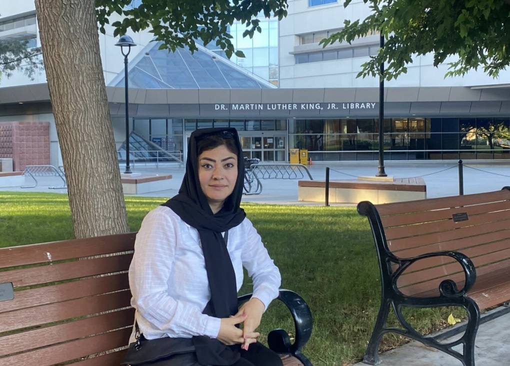 A woman with a black headscarf and white shirt sits in a bench in front of a library