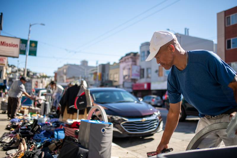 A man in a hat stands next to a variety of wares with houses in the mission in the background