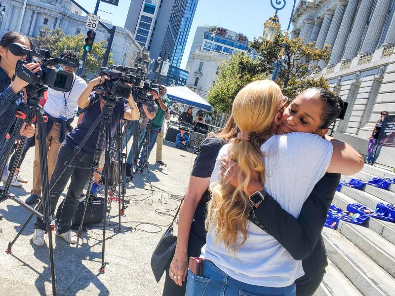 two women hug at a rally in front of city hall, with news cameras all around