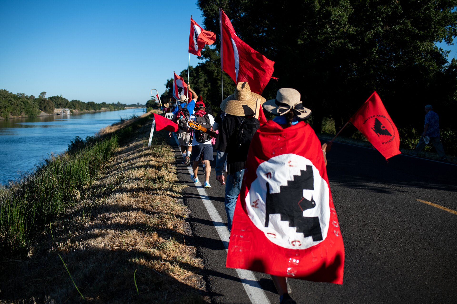 line or marchers, one draped in union flag, walk away from the camera along a rural highway next to a river