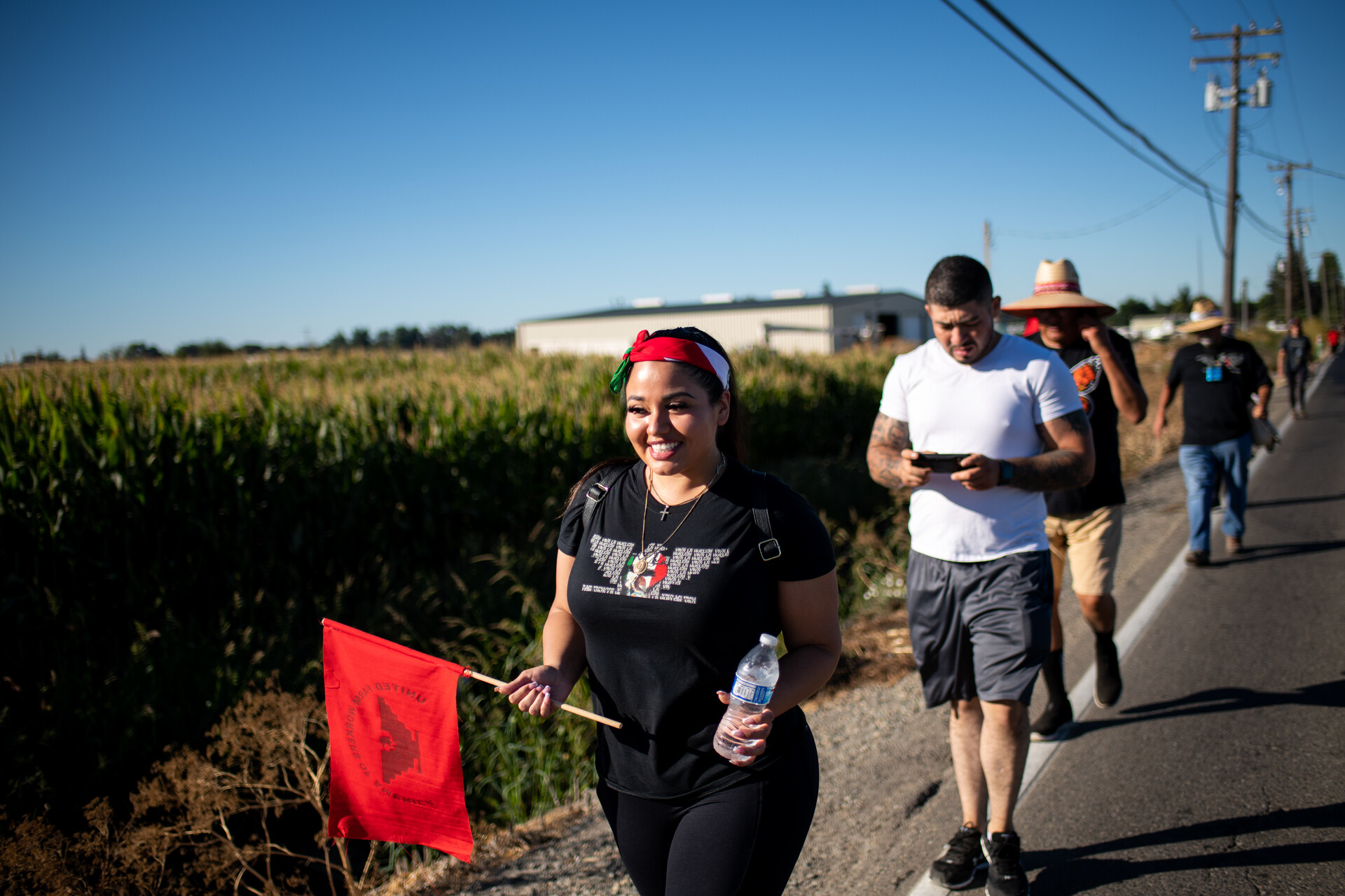 smiling woman holds red flag as she walks at front of line of marchers along rural road in the sunshine