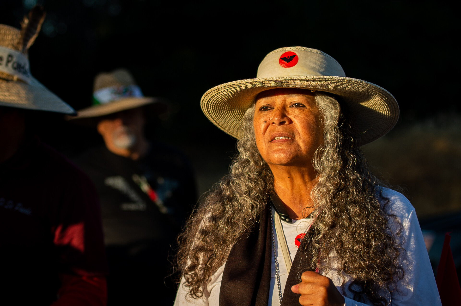 woman with long grey hair wearing hat and light by early morning sun speaks as others listen