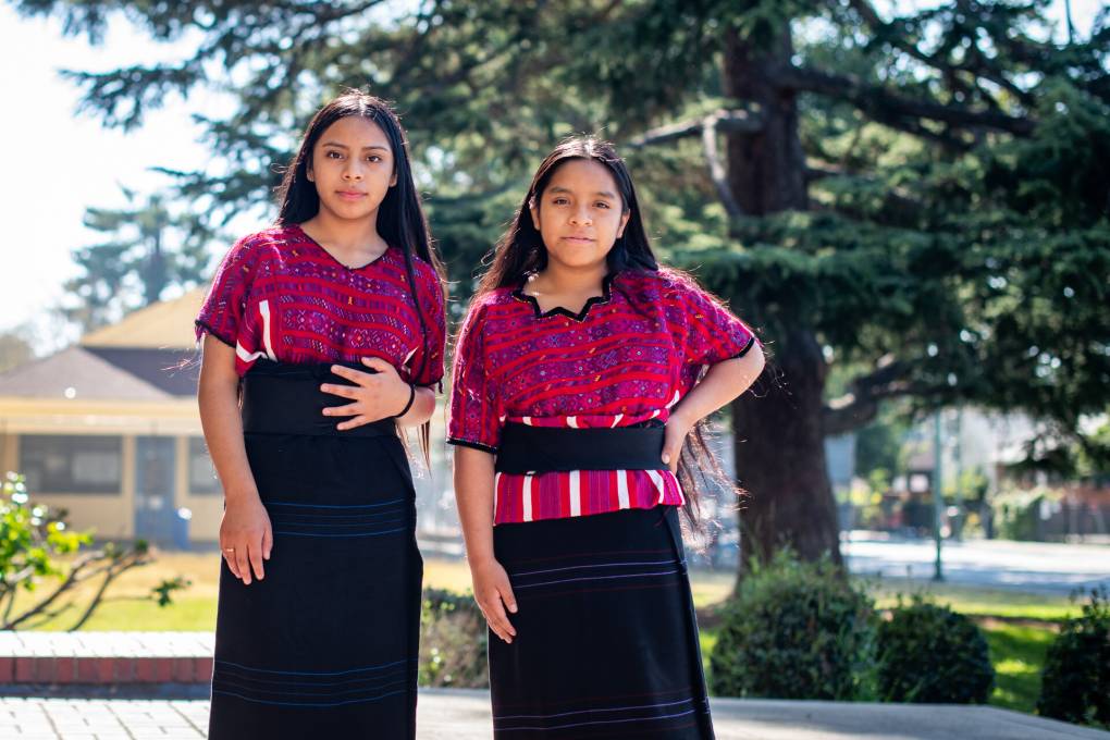 Two young girls wearing red decorative shirts and long black skirts stand outside.