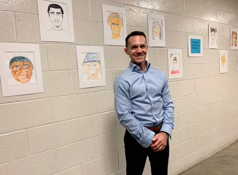smiling man in button up shirt stands in front of portrait drawings hung on a concrete interior hallway wall behind him