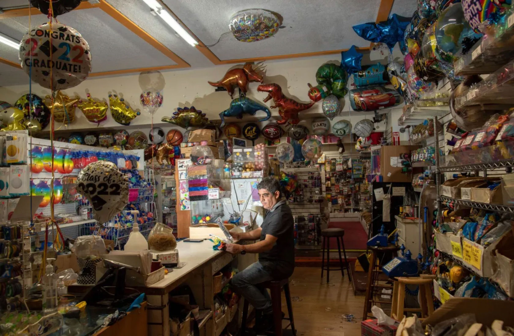 man sits at desk in store surrounded by cornucopia of inflated hearts, animals