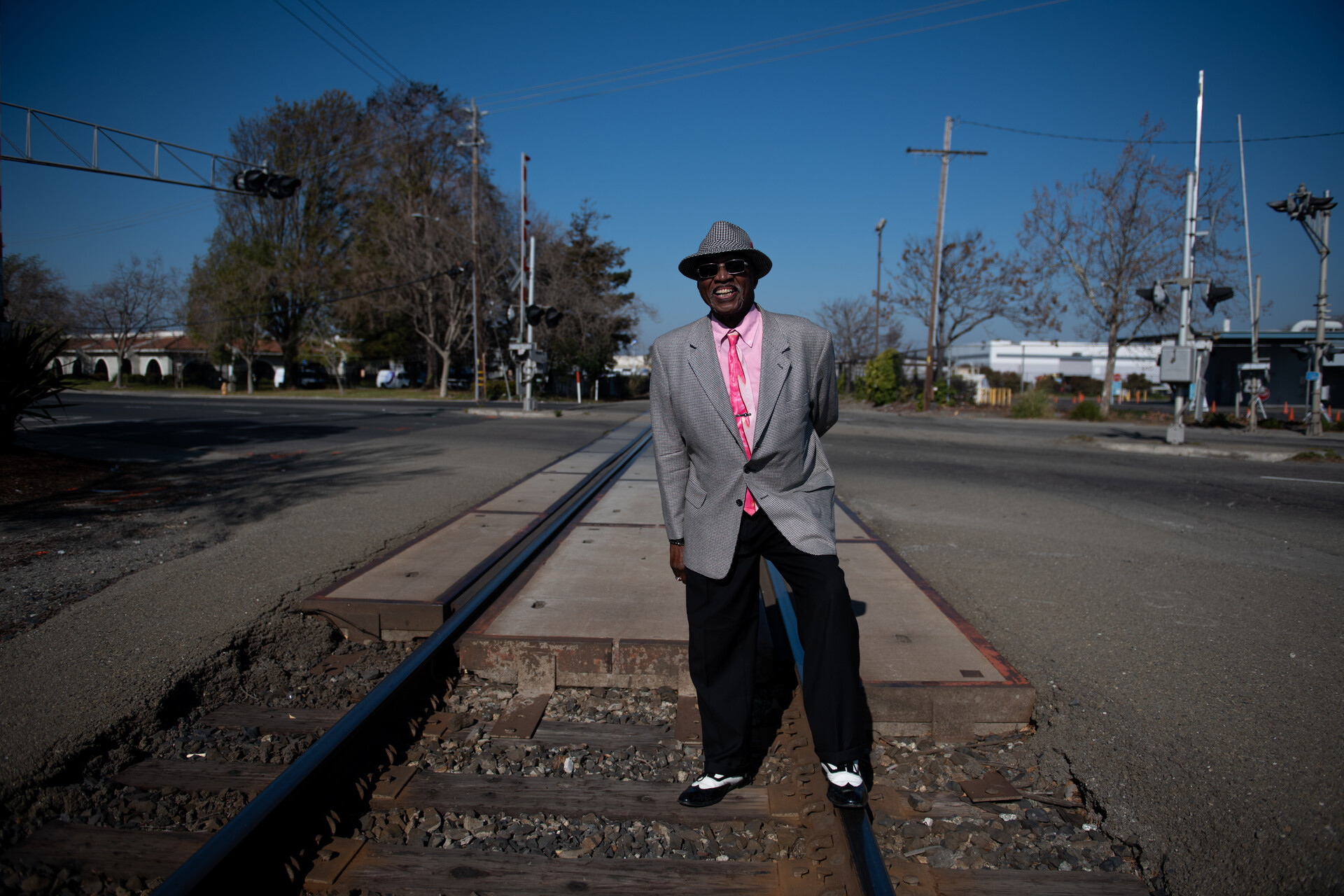 A Black man stands in the middle of the railroad tracks wearing a stylish suit jacket with pink shirt and tie, fedora, and black and white shoes.
