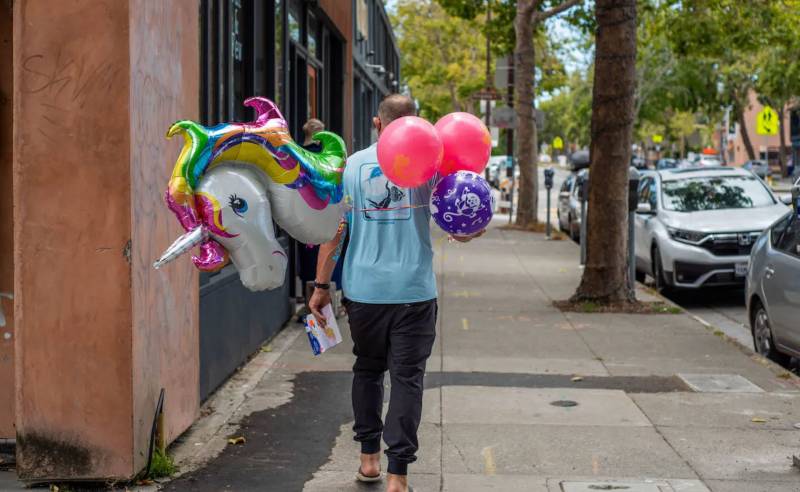 person walking away from camera on sunny day down sidewalk carrying large balloons including unicorn-shaped ones