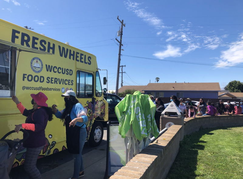 A yellow truck has a sign that reads "Fresh Wheels WCCUSD Food Services" outside with several people lined up waiting in masks.