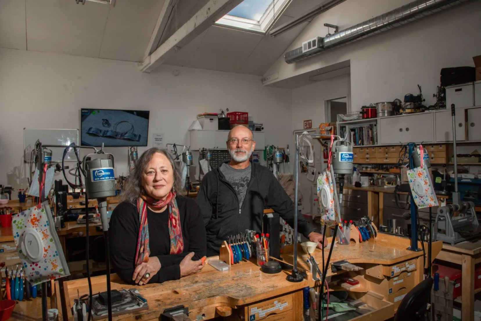 Couple stand behind worktable in well-lit workshop, facing camera