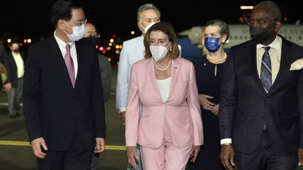 Nancy Pelosi, wearing a mask, walks in front of a group of officials.