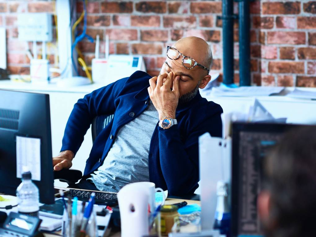 A man in a blue shirt and glasses sits in an office with his face to his hands in an expression of tiredness