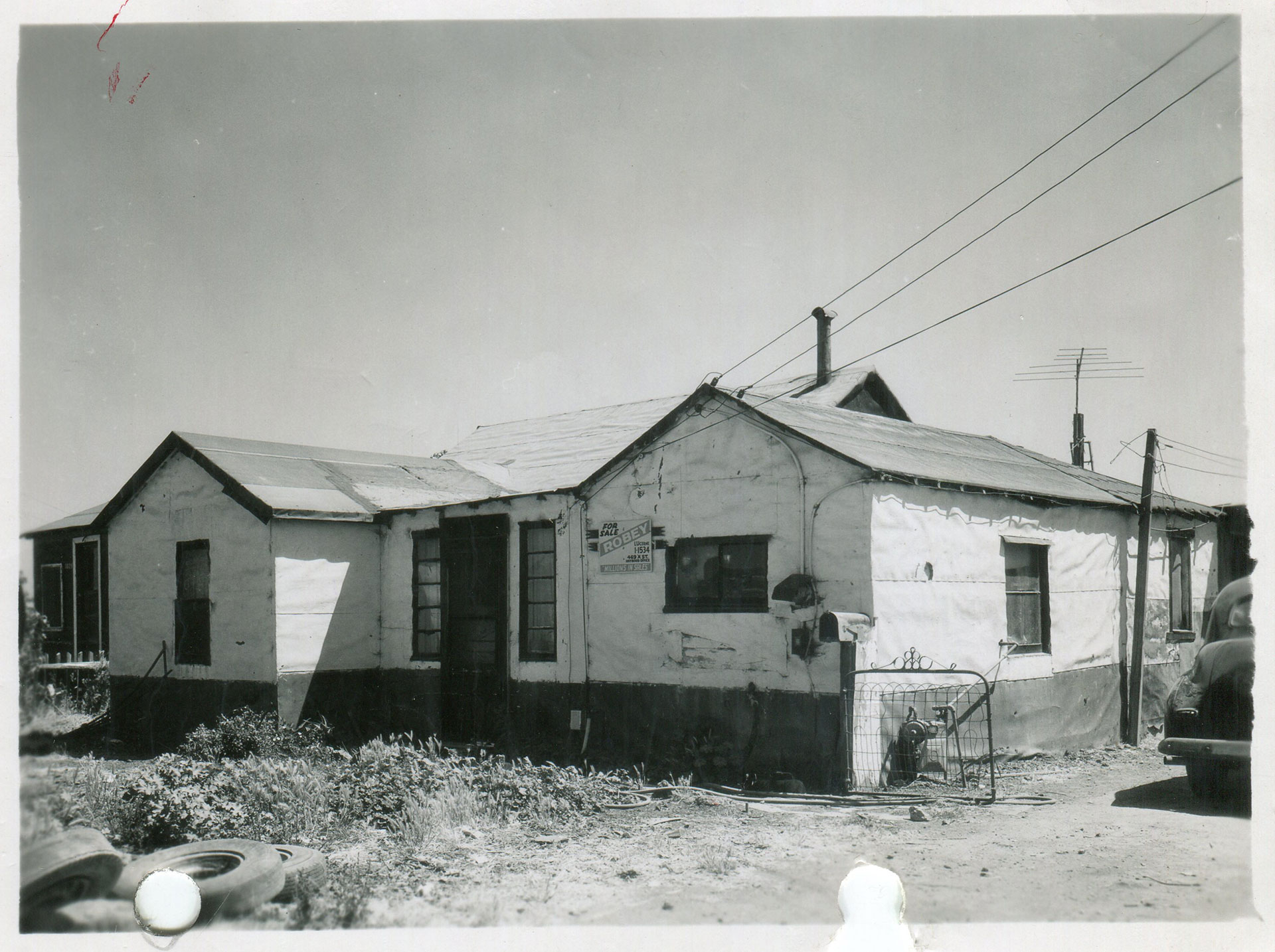 Black and white photo of a small house with paper siding, a pile of tires out front, a dusty yard and a for sale sign.