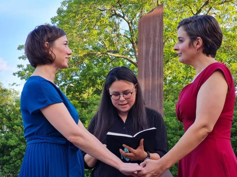 Two white-presenting women, Carlie Brown and Molly Pela hold hands and exchange wedding vows as their friend, Julie Takahashi, an Asian woman, officiates the ceremony in the middle of them. Both Carlie and Molly have short brown hair. Carlie is wearing a blue dress, and Molly is wearing a red dress. Julie is wearing a black shirt.
