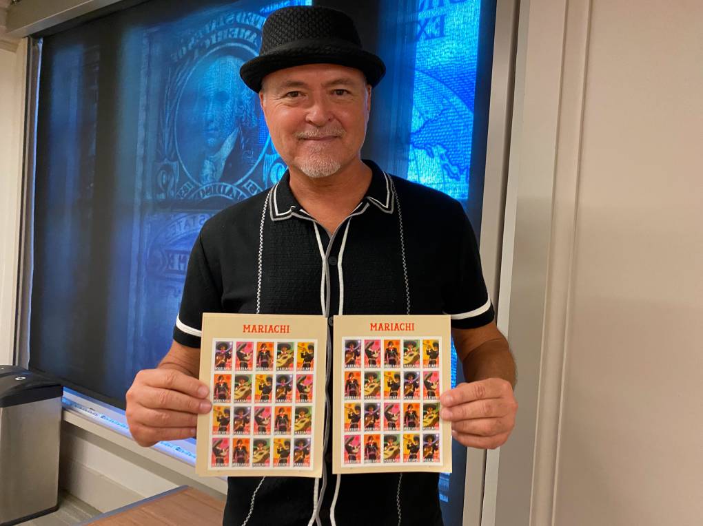 a man in a black hat holds up postage stamps that feature mariachi musicians