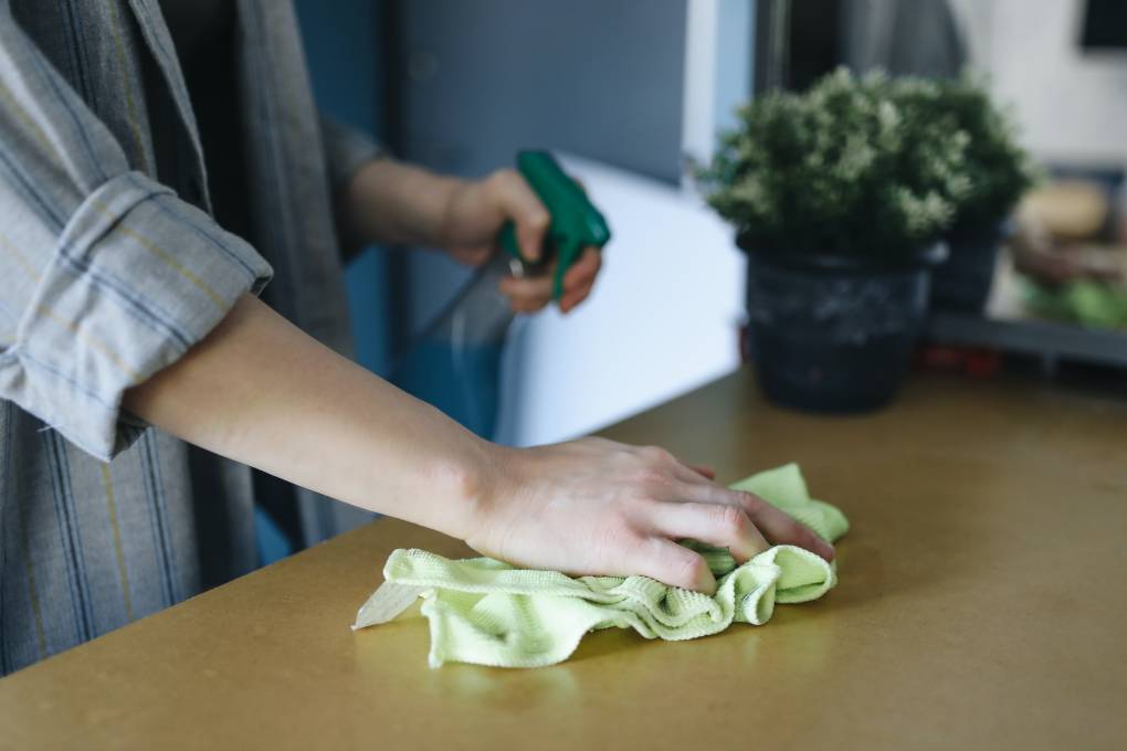 A person cleans a table with a rag in an indoor space and holds a spray nozzle with their other hand.
