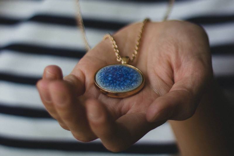 A hand with palm upturned holding a gold pendant with a blue stone. We can't see the face of the person, but they're wearing a black and white striped shirt.