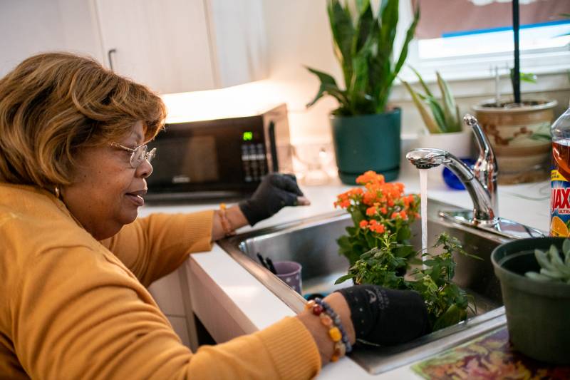 A woman wearing glasses, black gloves, bracelets and a yellow shirt rests her hands in front of a sink counter while water runs from a faucet into a plant.