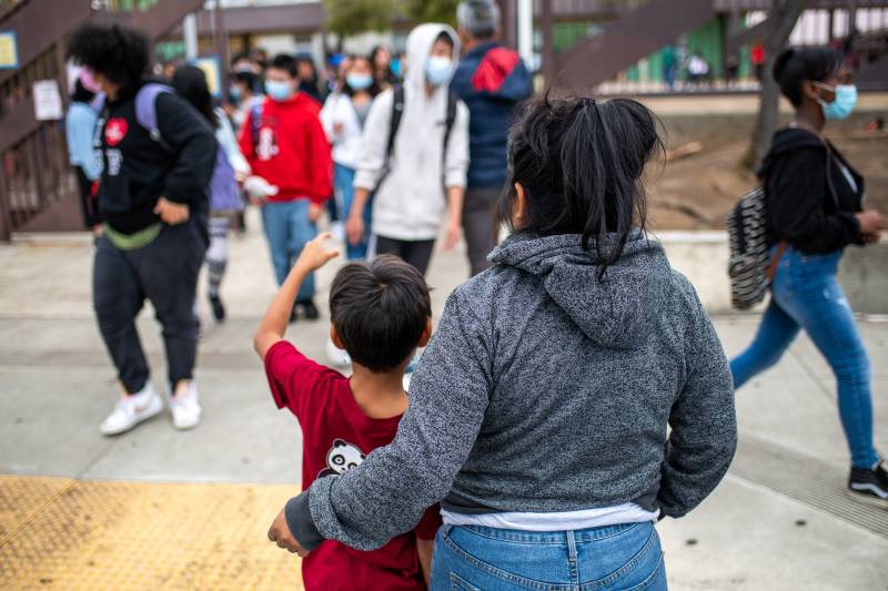 A Hispanic woman and her young son are seen from behind as high schoolers exit school.