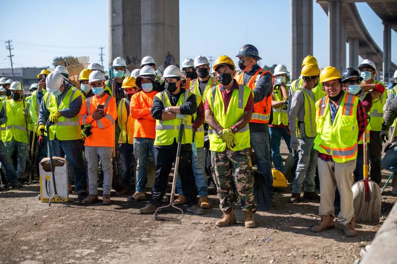Several people in hard hats and construction bests stand in a line under a freeway overpass