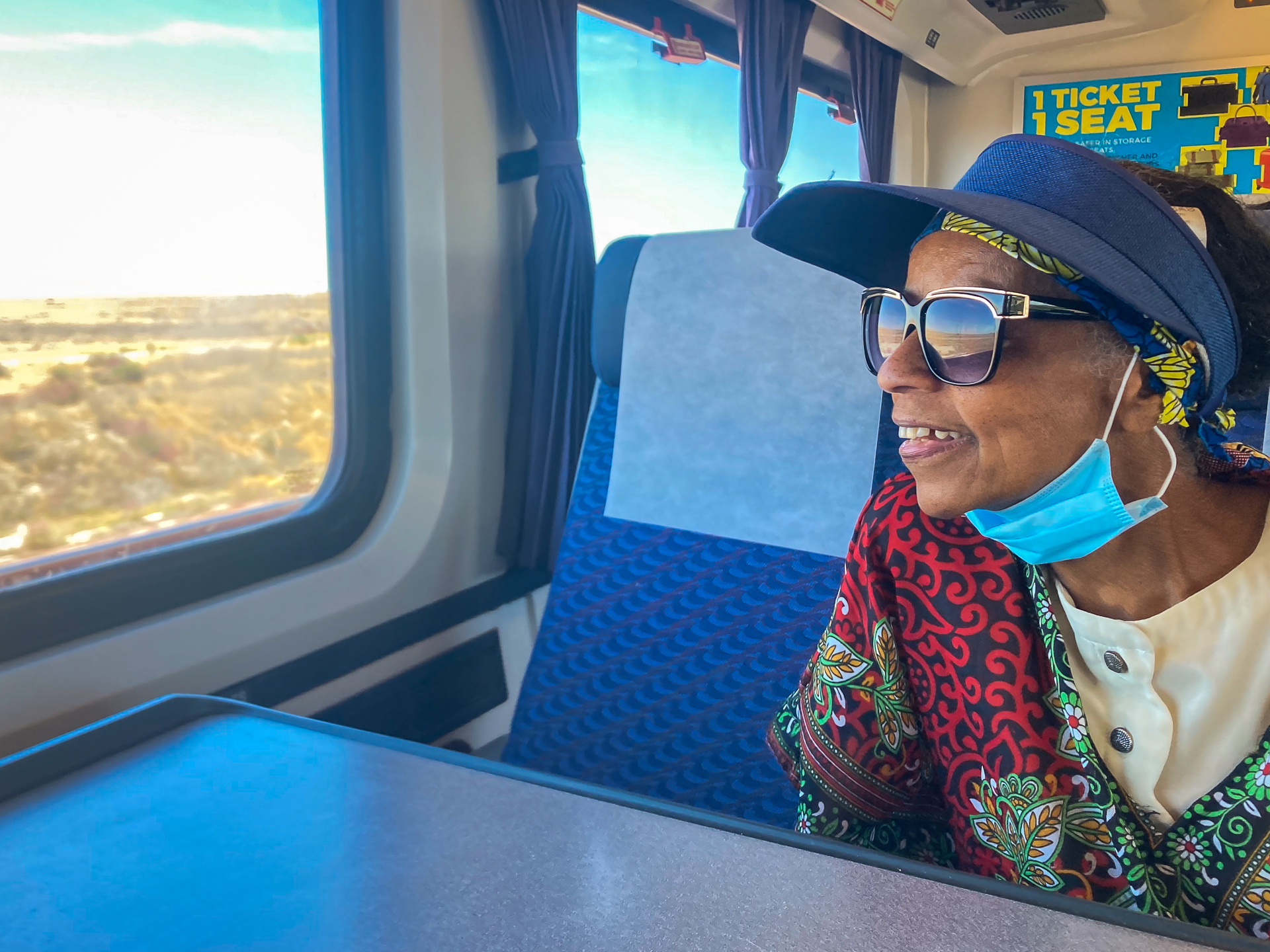 an older woman sits on a train wearing a hat and sunglasses and looking out the window