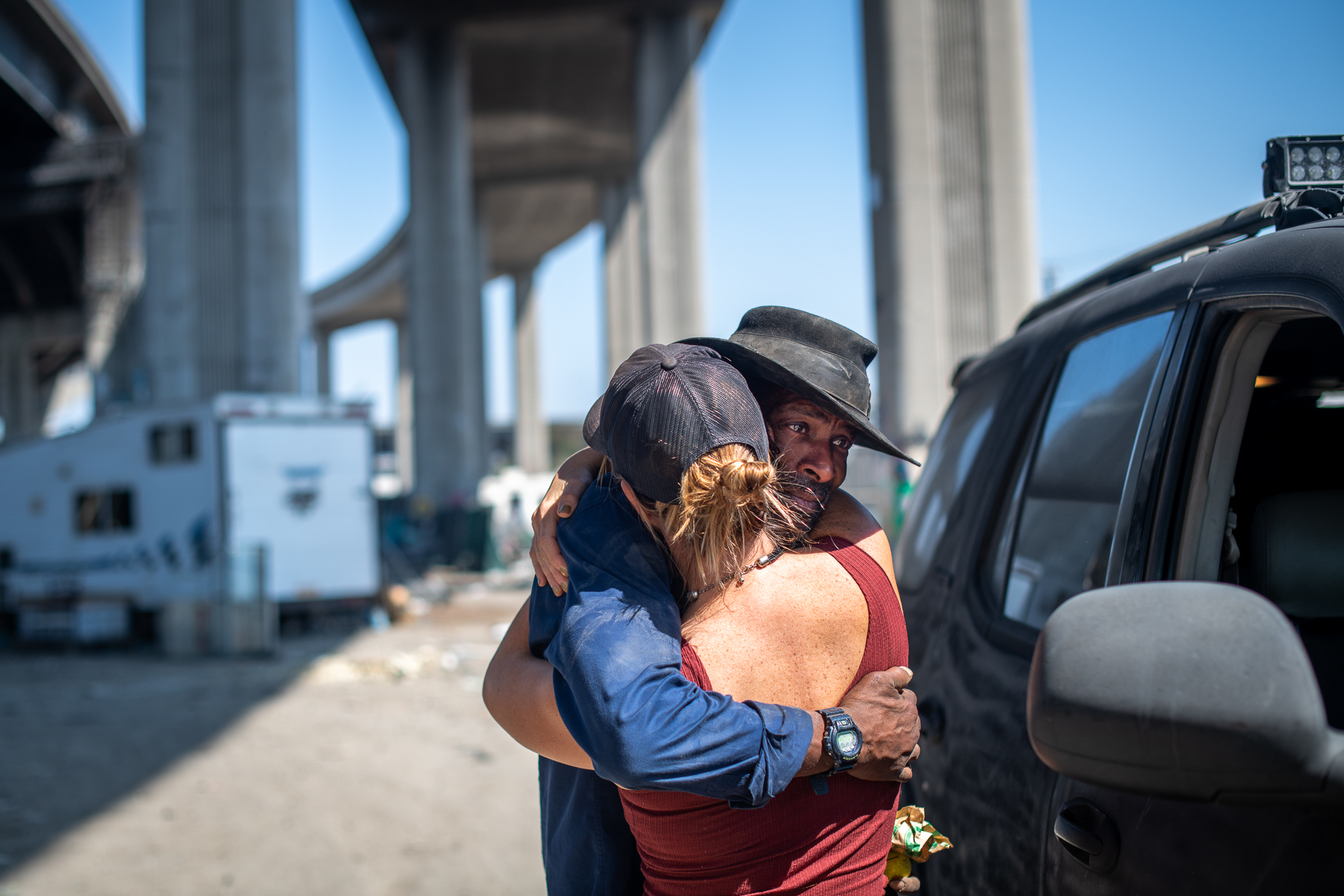 A Black man is photographed hugging another person, with his face visible over their shoulder. He is wearing a black wide-brimmed hat and blue shirt, and his friend (whose face we can't see) is wearing a red vest and a black baseball cap. They are photographed under a freeway overpass.