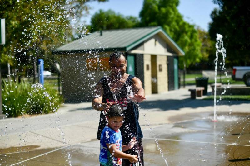 A man and a boy stand in a sprinkler at a park.