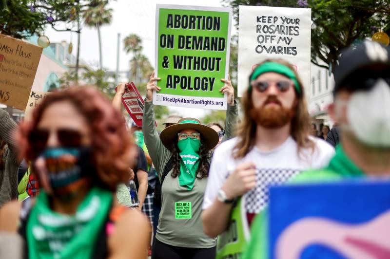 A crowd of protestors face the camera, but all are out of focus except for one person in the middle of the frame and the crowd. That person, sharp and in focus, wears a grey top and a green bandana and holds a sign above their head that reads "Abortion on demand and without apology."