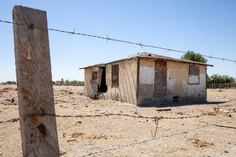 A sagging, abandoned wooden building sits in dry soil behind a barbed wire fence.