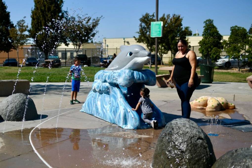 Two children play in a playground sprinkler, with a dolphin-water cave, as a woman looks on.