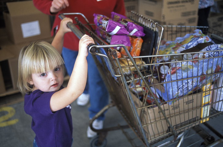 A small blonde-haired girl pushes a shopping cart full of groceries at a foodbank next to her mother.