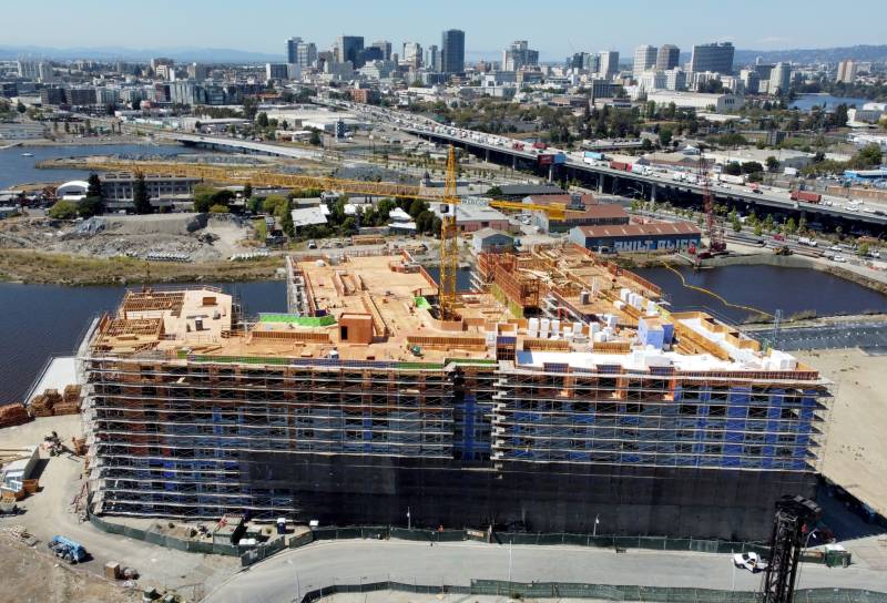 An aerial view of a housing development in Oakland. The building is more than a frame but not yet complete, and a crane sticks out from the roof. Far in the background, Oakland's downtown area.