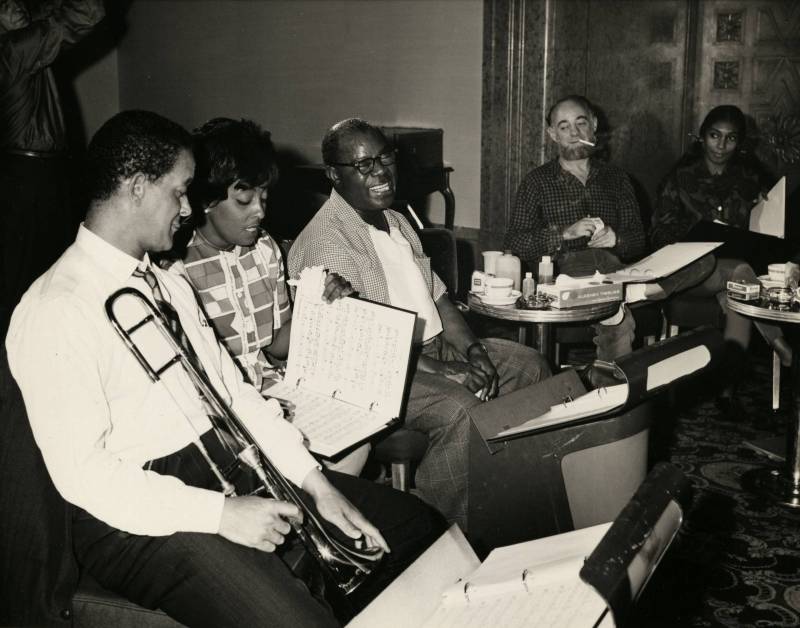 black and white photo of a group of jazz legends seated, smiling, looking at sheet music while one man holds a trombone