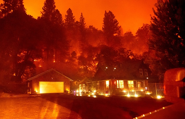A house is surrounded by fire with trees around it glowing red and a hazy orange sky above.
