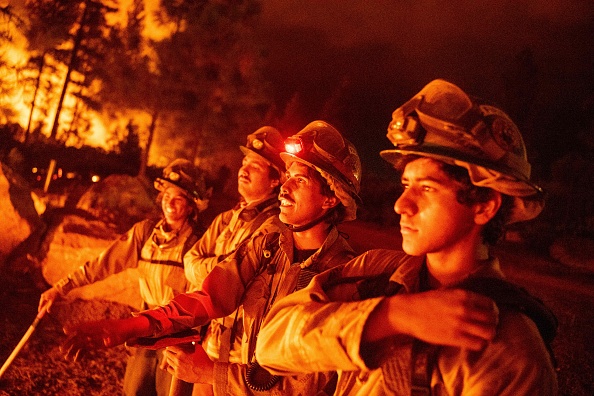Four firefighters, standing shoulder to shoulder, faces glowing orange, look out onto an ongoing blaze with burning trees in the background.