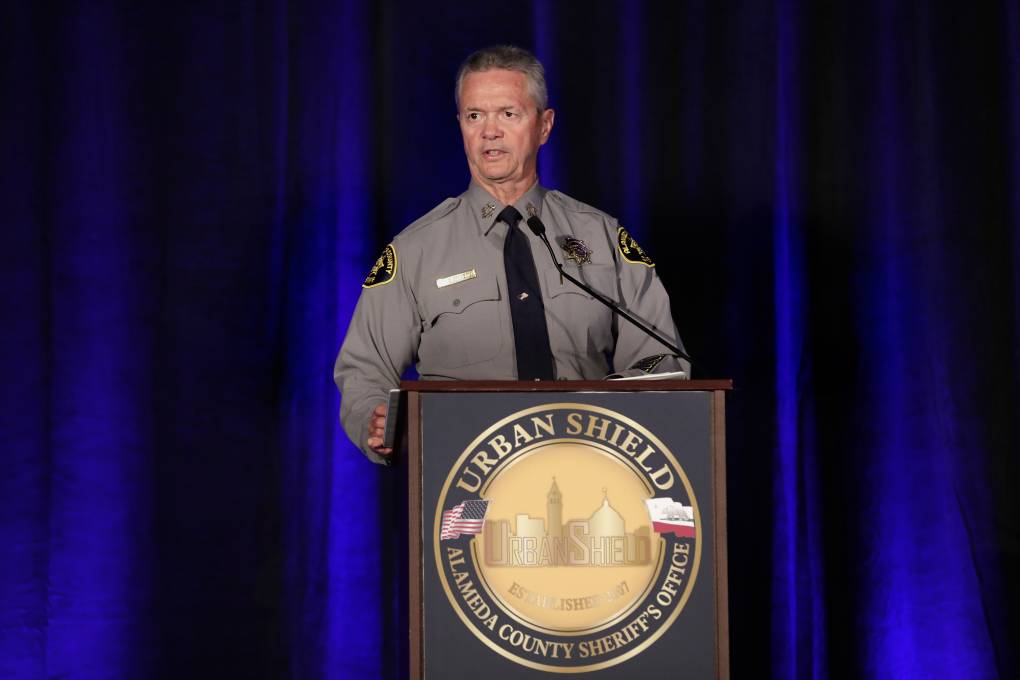 a sheriff in a grey uniform speaks on a stage in front of a blue curtain