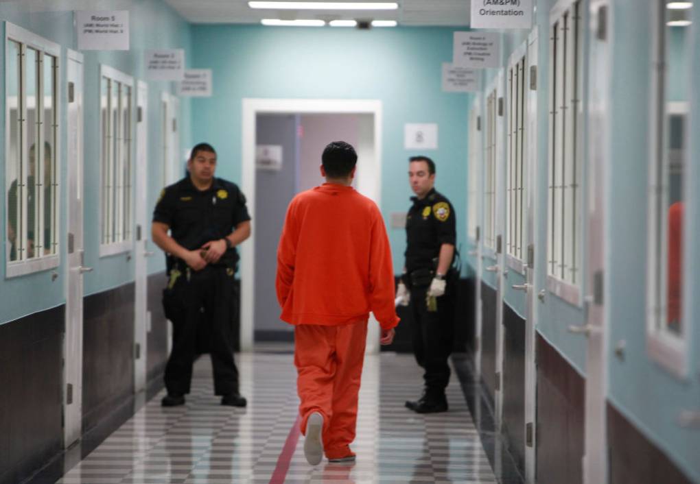 A man dressed in an orange jumpsuit walks towards two men dressed in correctional officer uniforms in a facility.