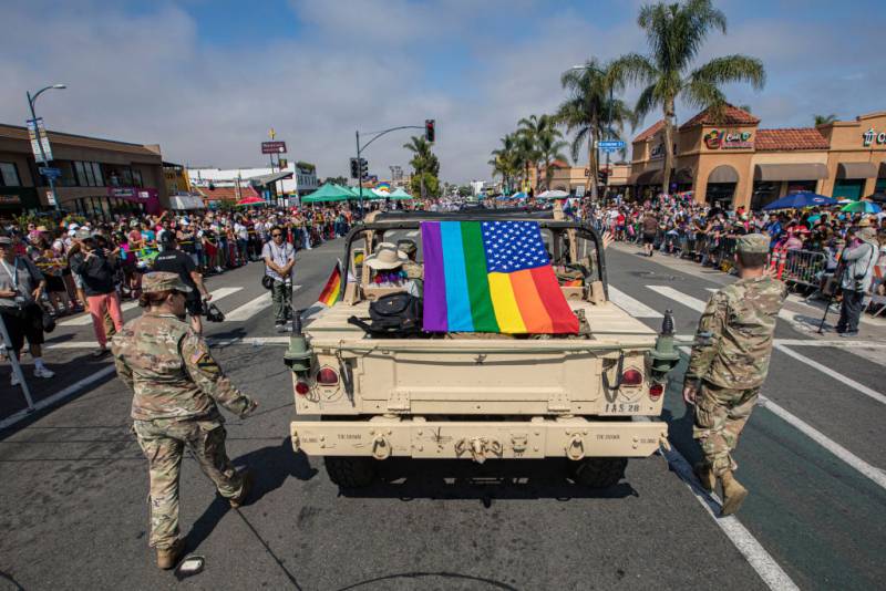 Two people dressed in military clothing walk on both sides of a military vehicle with a rainbow flag in the middle with thousands of people lining the sides of the street.