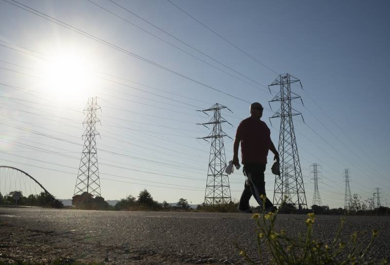 A bright white starburst of sun shines in a pale blue sky through dozens of electricity lines. Electrical towers line the bare ground. A man, backlit by the sun and appearing as a silhouette, walks in front of the towers, toward the right of the image.