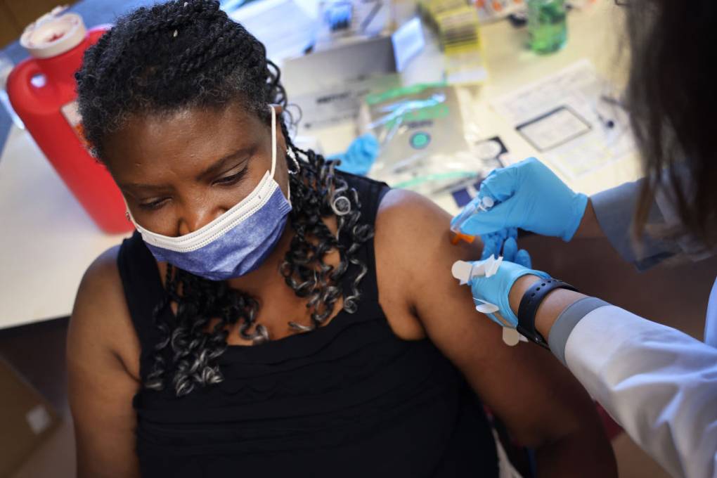 A woman wearing a face mask and a black sleeveless shirt sits as hands wearing blue gloves inject a syringe into her arm while holding the arm in place.