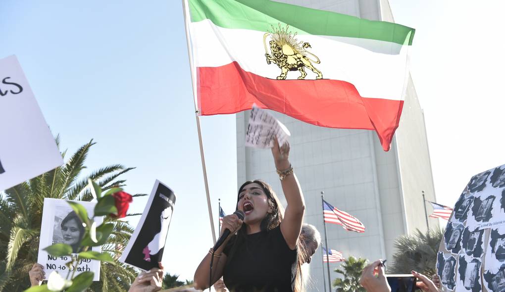 A woman appears to shout into a microphone with her other arm held up. Behind her, an Iranian flag flows in the wind.