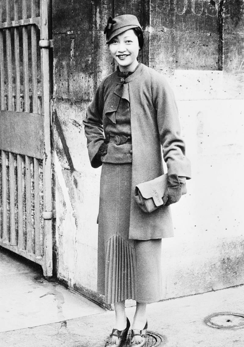 A black-and-white photo of a Chinese-American woman standing in front of a hotel wall in Paris in 1935. She's wearing a narrow skirt with kick-pleats, a belted jacket, a cloche hat, and tasseled shoes with heels. She's holding a clutch bag and smiling with delight.