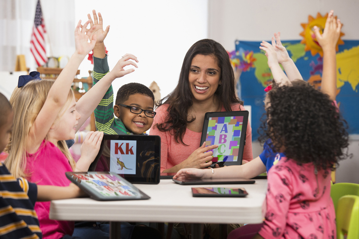 A teacher of South Asian descent points at a tablet with letters on it as smiling children of different races raise their hands