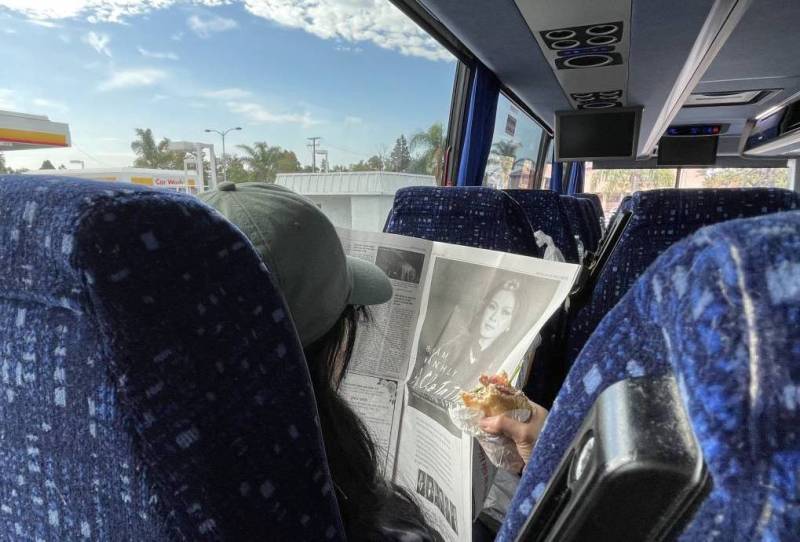 A person wearing a hat holds a half-eaten sandwich and a newspaper in a bus seat.